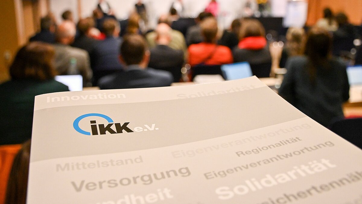 Pressemappe des IKK e.V.  im Vordergrund, im Hintergrund Menschen auf einer Tagung
