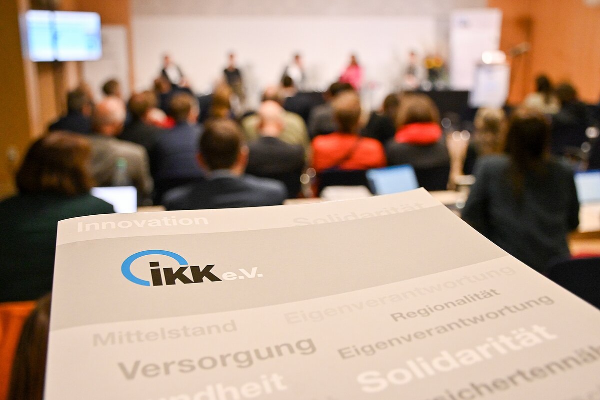 Pressemappe des IKK e.V. im Vordergrund, im Hintergrund Menschen auf einer Tagung
