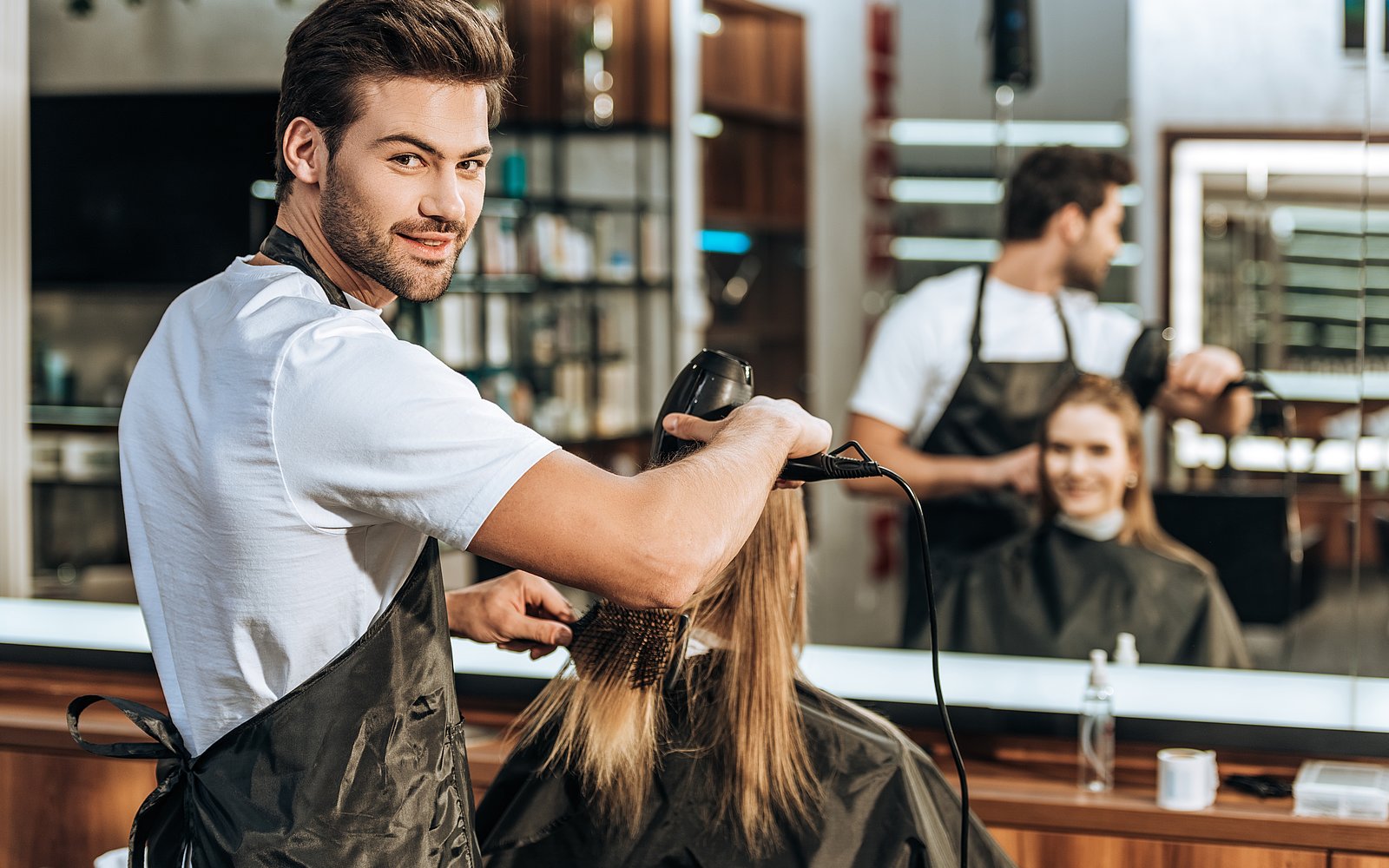 Friseur föhnt einer Frau die Haare und gruckt dabei in die Kamera