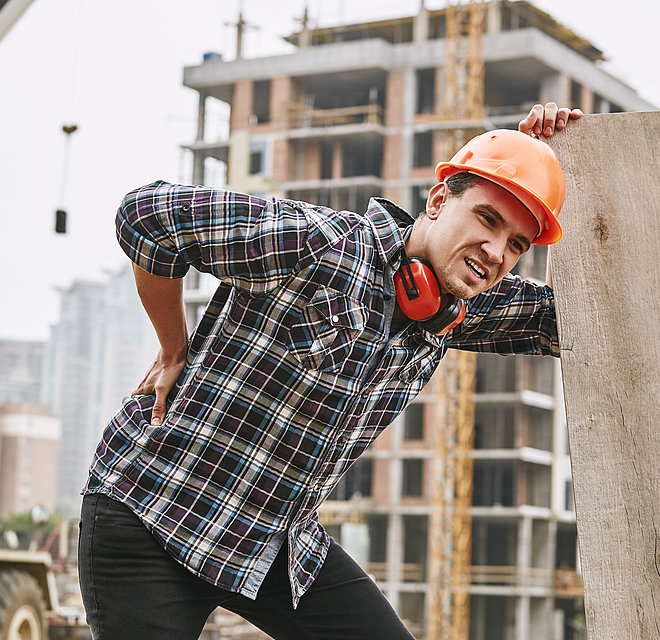  Bauarbeiter im Schutzhelm mit Rückenschmerzen bei der Arbeit auf der Baustelle.